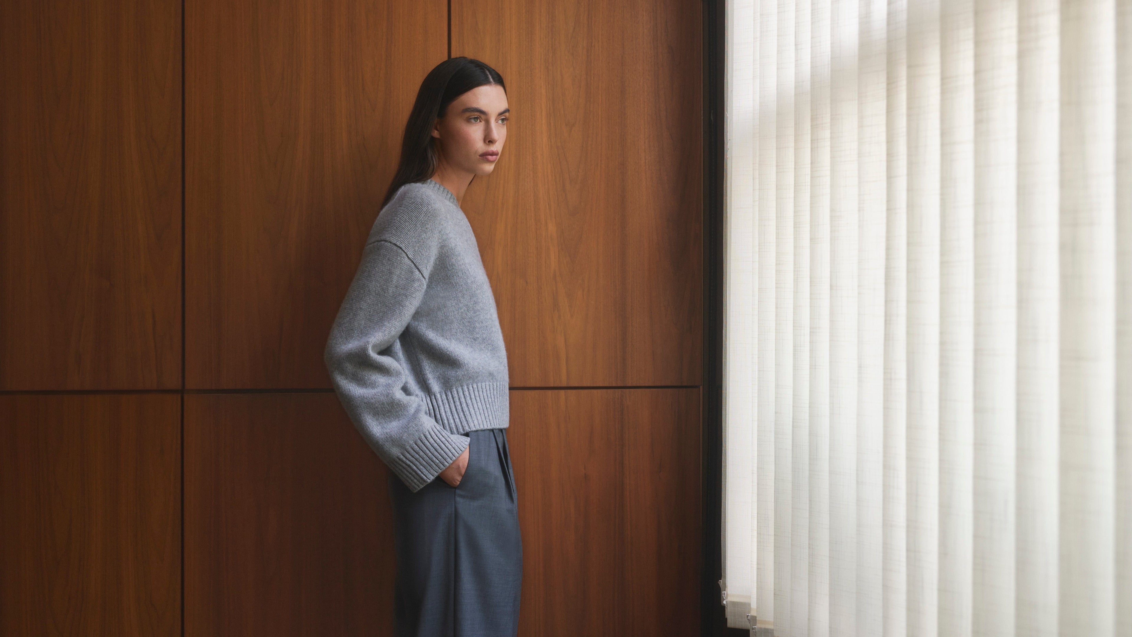 Woman in a light gray sweater and pants standing against a wooden wall with a window with blinds. Pringle of Scotland Women's Cashmere