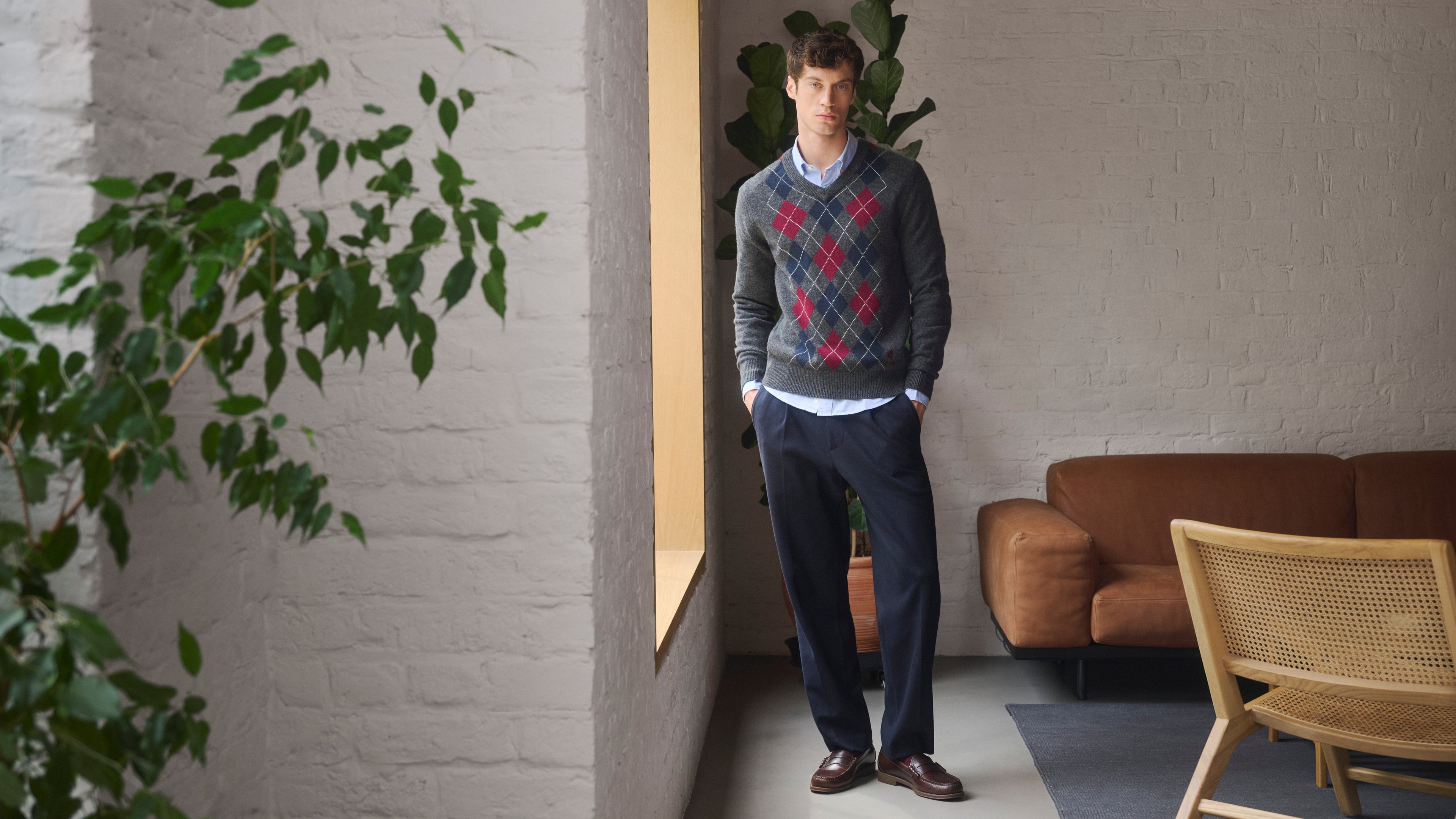Man wearing classic argyle sweater by Pringle of Scotland standing in a room with plants and furniture.