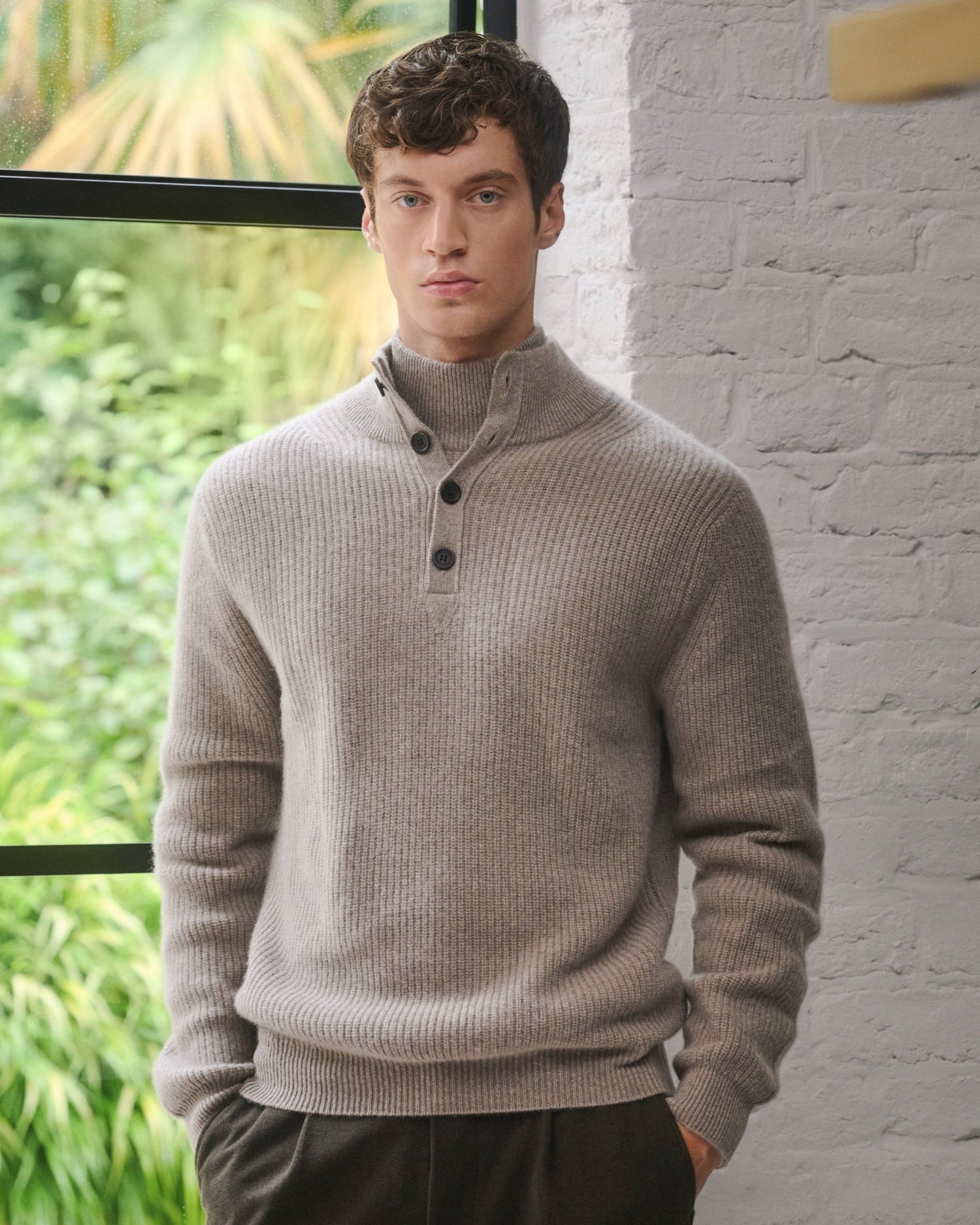 Man wearing a beige Pringle of Scotland cashmere sweater standing against a white brick wall with greenery in the background
