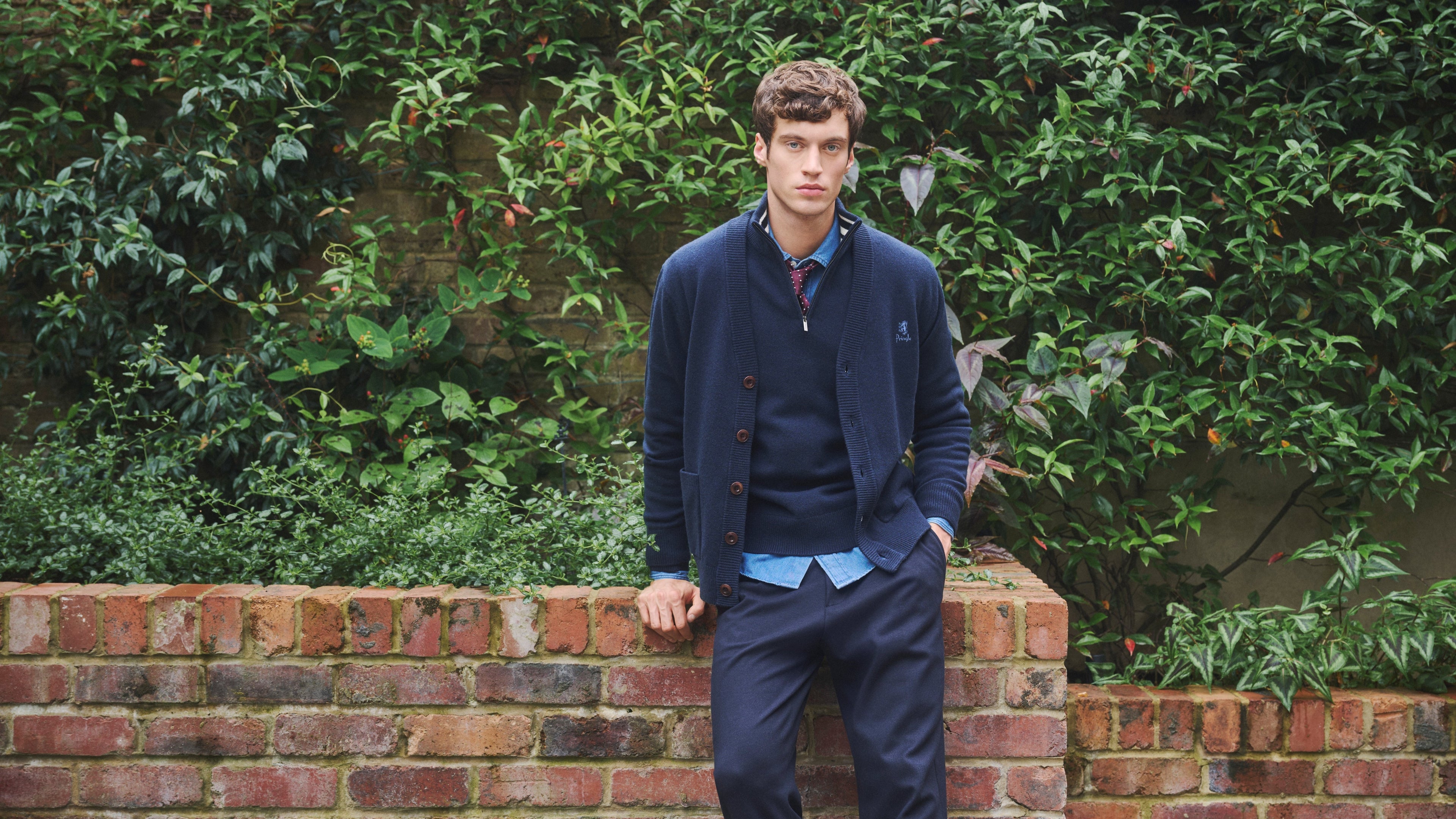 Man standing against a brick wall wearing a Pringle of Scotland cardigan with green foliage in the background