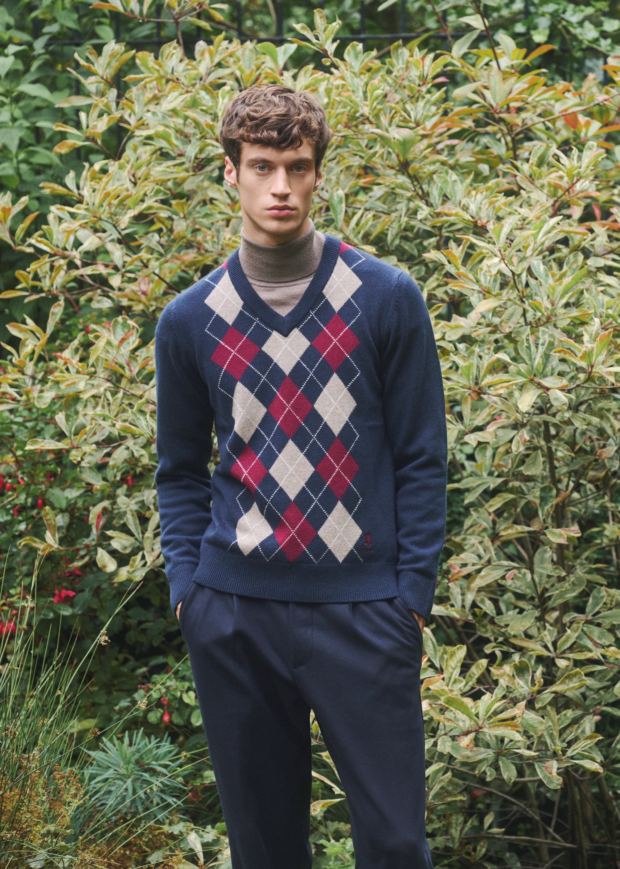 Man wearing a blue argyle sweater  by Pringle of Scotland standing in front of green foliage