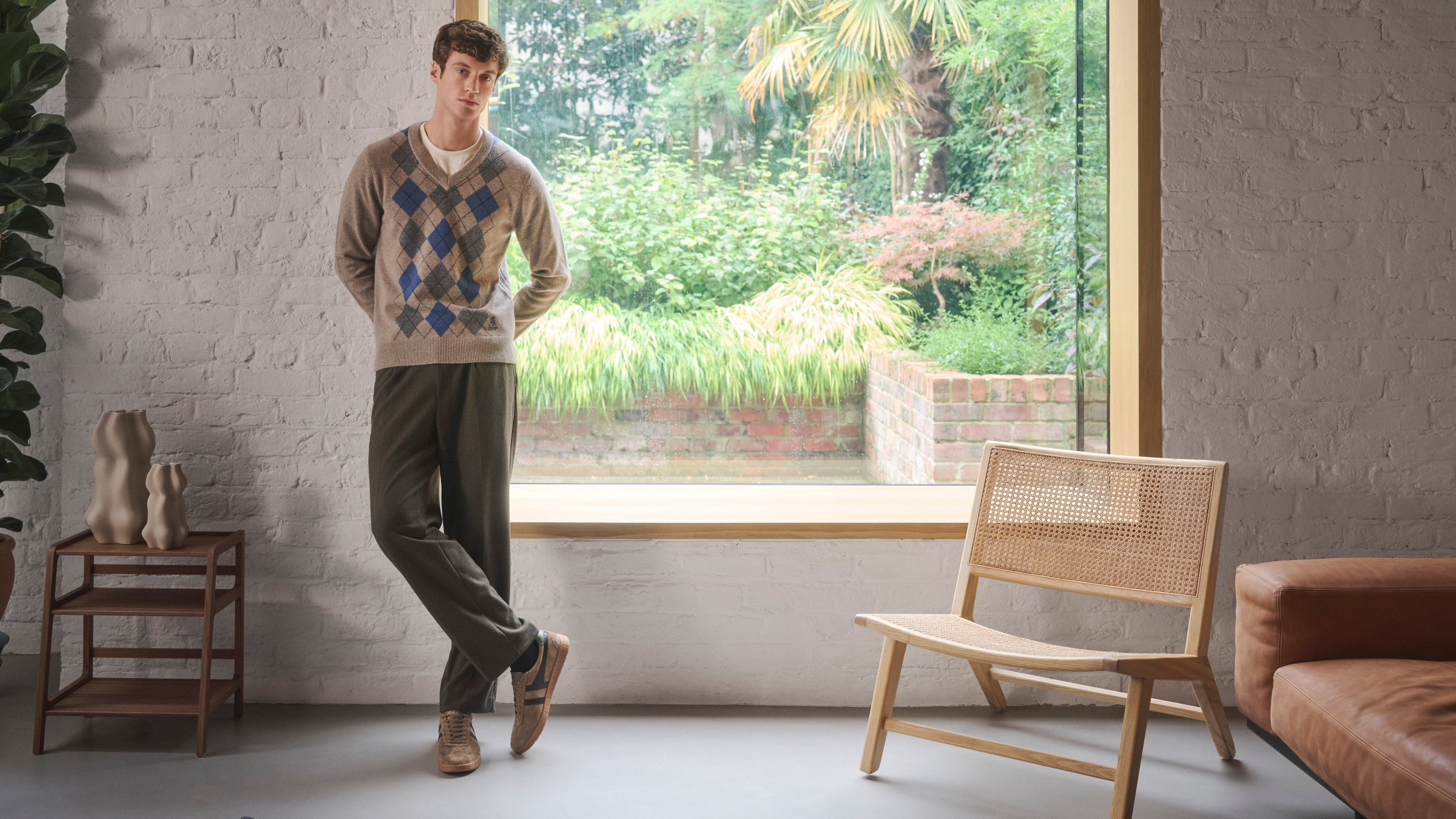Man standing in a modern living room with a large window, garden view, and minimal decor wearing Pringle of Scotnad argyle jumper