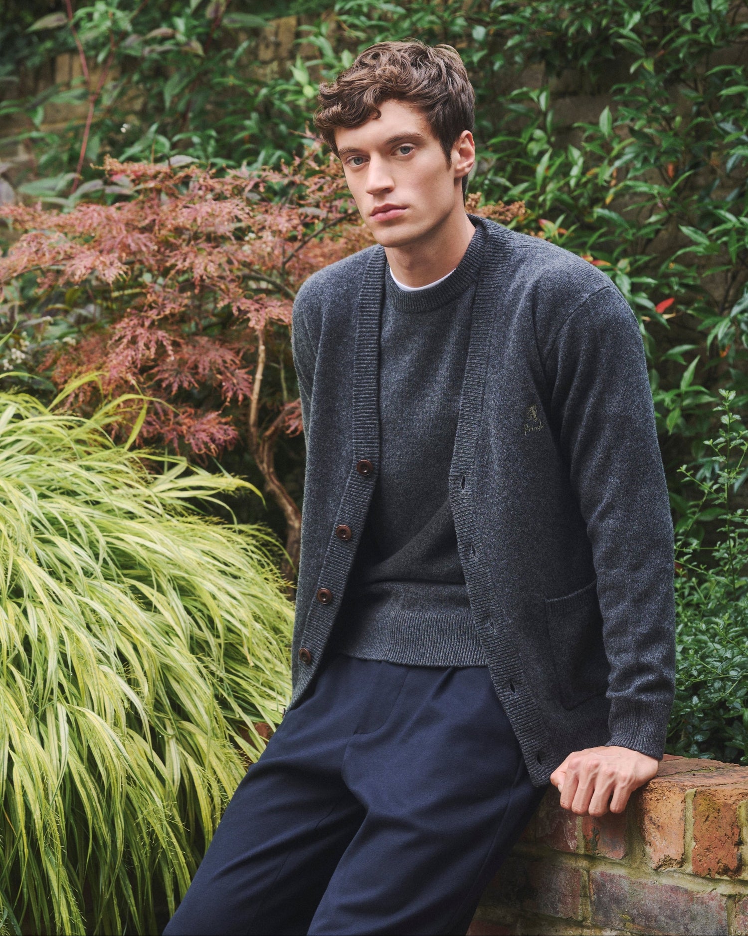 Man sitting on a brick wall in a garden surrounded by greenery wearing Pringle of Scotland Supersoft Lambswool Knitwear in Charcoal