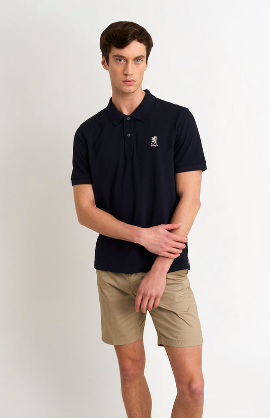 Man wearing a navy polo shirt with a logo and beige shorts on a white background
