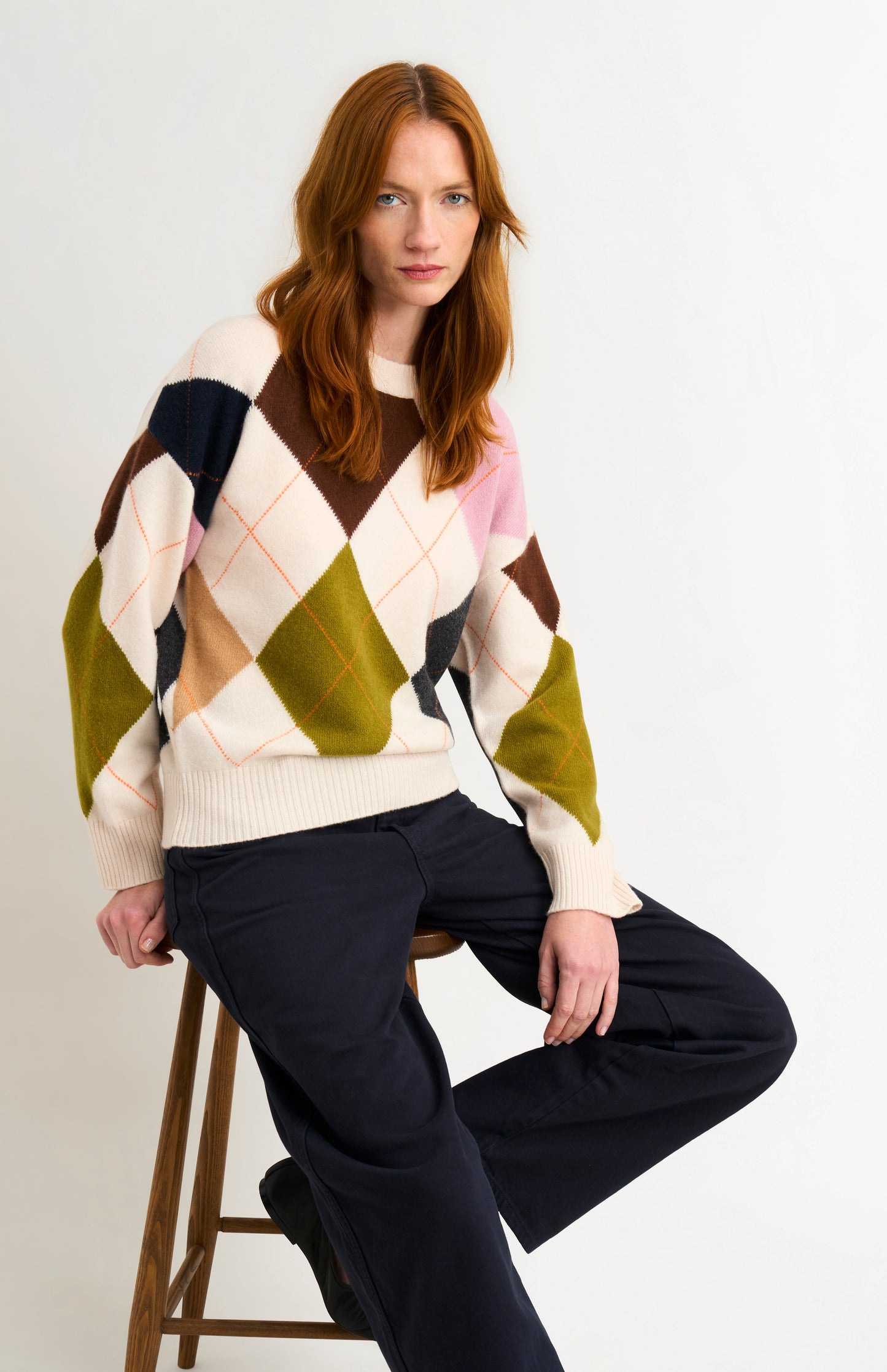 Woman wearing a colorful argyle sweater sitting on a stool against a white background