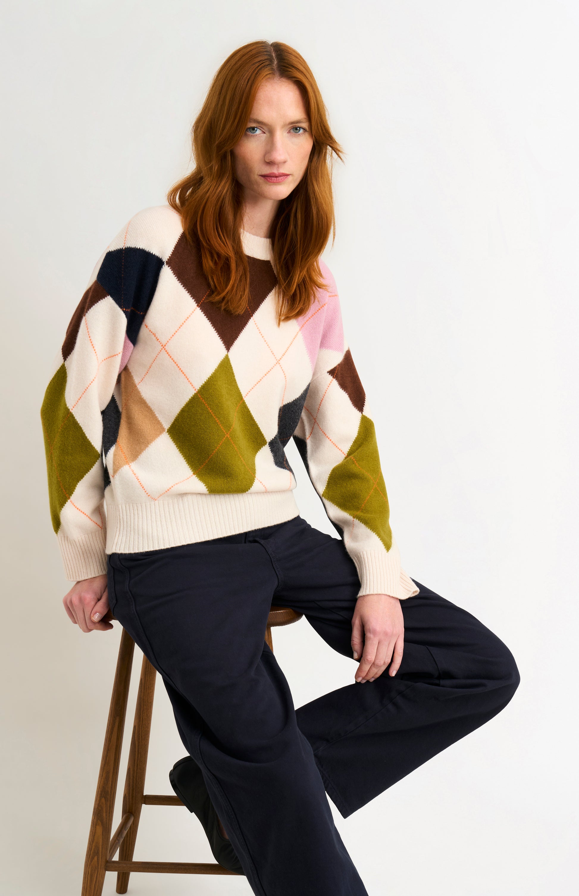 Woman wearing a colorful argyle sweater sitting on a stool against a white background