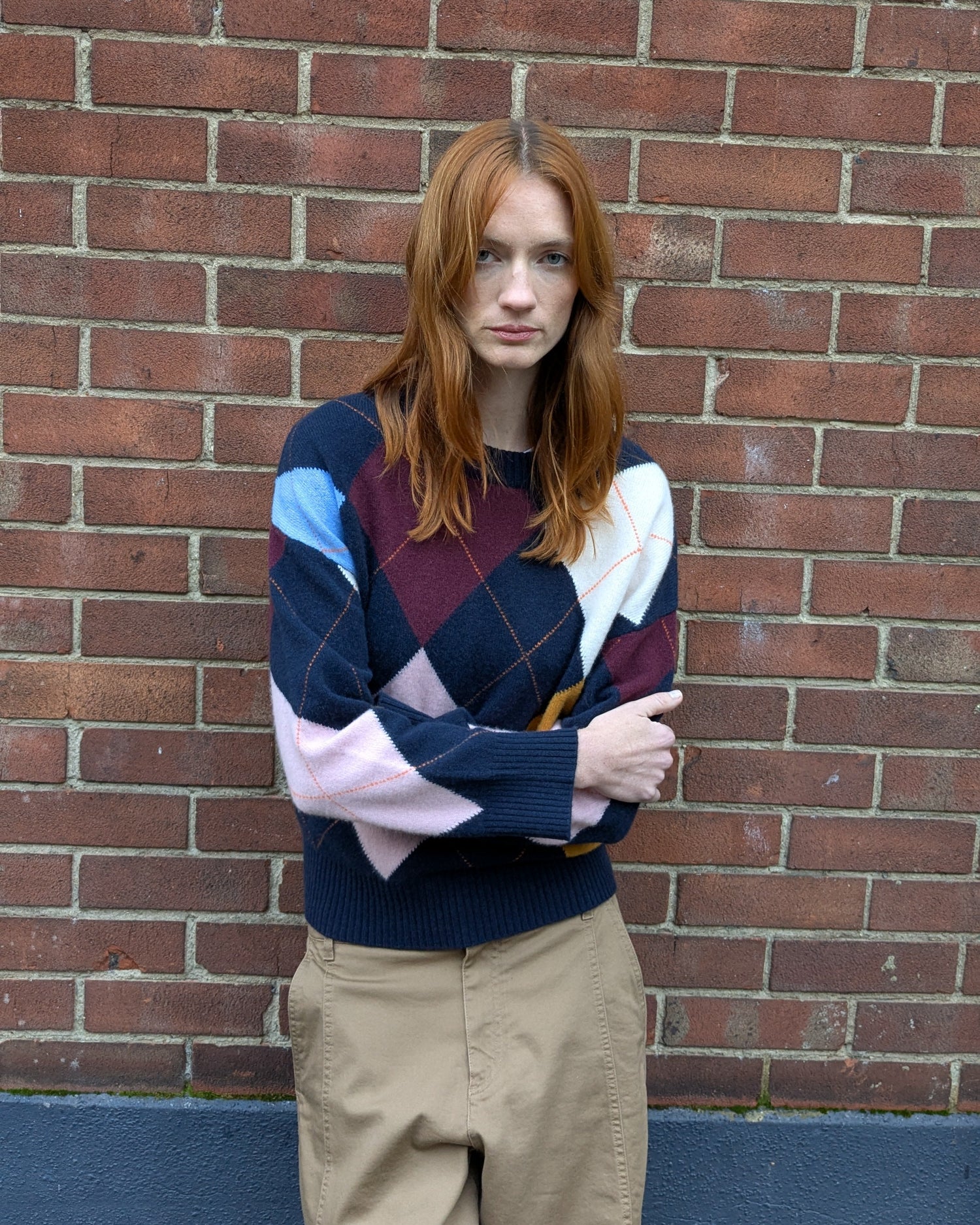 Person wearing a colorful sweater and beige pants against a brick wall.
